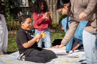 Kinderen maken samen een kunstwerk op een groot doek in het gras in Amsterdam-Zuidoost. Foto: Joni Reiche.