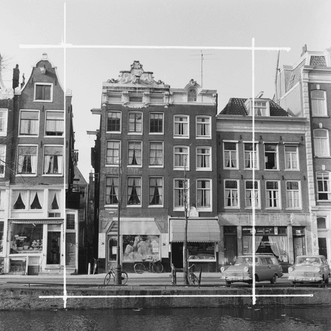 Zwart-witfoto van huizen aan een Amsterdamse gracht met geparkeerde auto's en fietsen.