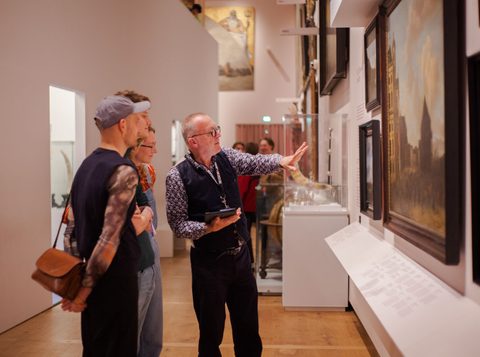 drie mensen worden rondgeleid door panorama amsterdam. De rondleider wijst iets aan op een schilderij terwijl hij erover vertelt.