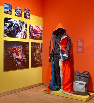 Beeld uit de tentoonstelling met een pop in een sheltersuit en foto's op de achtergrond.